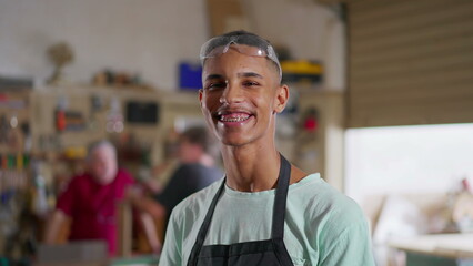 One young Brazilian employee of small business wearing uniform and protective eye-wear standing in workshop turning head to camera smiling
