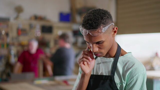 One young Brazilian employee of small business wearing uniform and protective eye-wear standing in workshop turning head to camera smiling