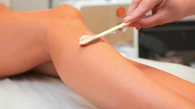 Woman specialist smears wax on stick on female leg in salon light office. Concept of epilation and equipment for procedure by professional