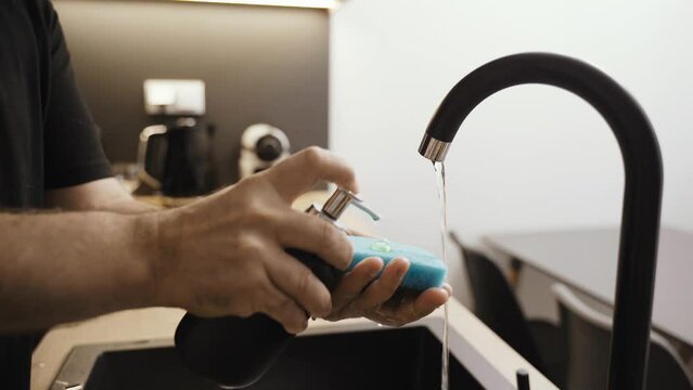 A man applies dishwashing detergent to a sponge, washes a white plate under running water. Close-up.