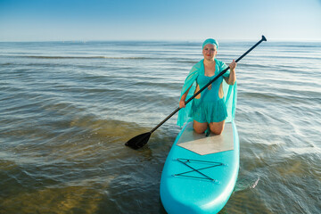 A Jewish woman in a turquoise bathing suit with a skirt and a headscarf on her knees on a SUP board sails away from the seashore.