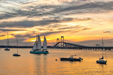 Sailboats at rest in Newport Rhode Island harbor as a peaceful sun sets behind the Newport bridge - Powered by Adobe