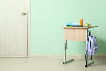 Modern school desk with backpack, bottle of juice and stationery in room near green wall