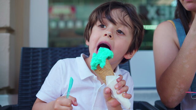 Child enjoying ice-cream cone at parlo shop during summer day. Mother brings cone closer to small boy's mouth to prevent from staining clothes
