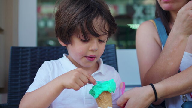 Child enjoying ice-cream cone at parlo shop during summer day. Mother brings cone closer to small boy's mouth to prevent from staining clothes