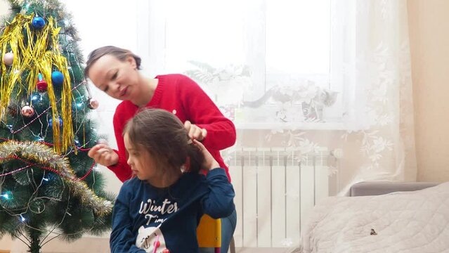 Mother braids her daughter's hair. Morning Christmas calmness relax cozy. New Year's decor in the bedroom. Xmas mood. Weekend leisure togetherness. Motherhood childhood. red sweater