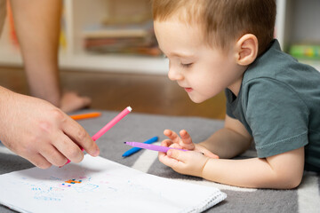 A beautiful little toddler boy of two or three years old with dad draws with markers in the album in the children's room at home sitting on the floor. Spending time with children. Selective focus