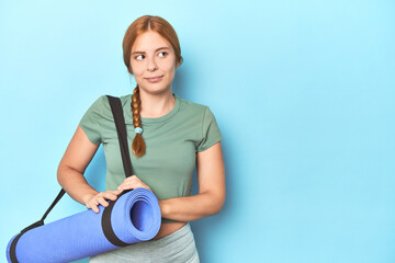 Redhead young woman holding yoga mat in studio dreaming of achieving goals and purposes