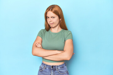 Obraz premium Redhead young woman on blue background frowning face in displeasure, keeps arms folded.