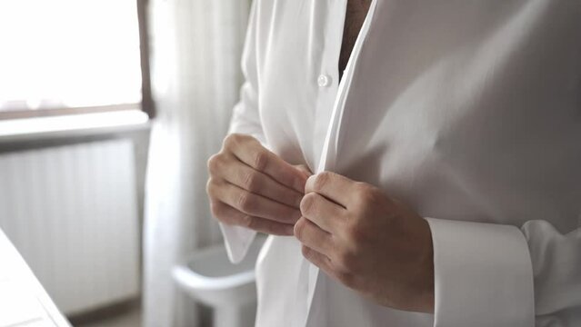 Man closes his shirt buttons. Young groom wears shirt and prepares for wedding. Elegance and slim model shirt. Close up of hands fastening buttons of elegant shirt. FullHD in slow motion.