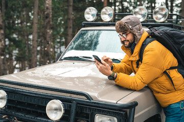 Young handsome hiker using mobile phone next to a jeep in the forest. Smiling man taking a break...