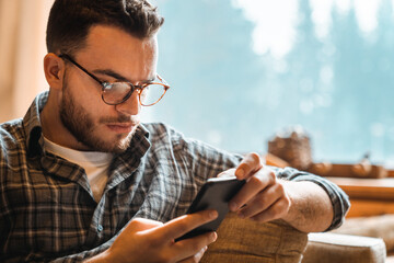 Close-up photo of focused male person wearing glasses and using mobile phone while sitting indoors...