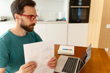 Obraz premium Serious attractive bearded man wearing red glasses, casual green t-shirt sitting at workplace