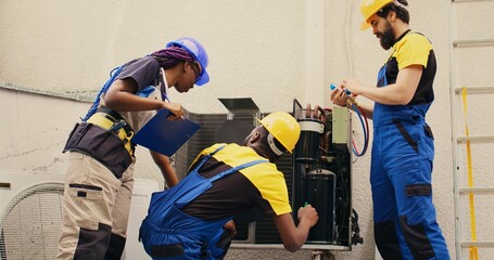 Expert repairmen cleaning layer of dirt and dust from air conditioner compressor coils while coworker uses manifold indicators to check for refrigerant leaks and read pressure in hvac system