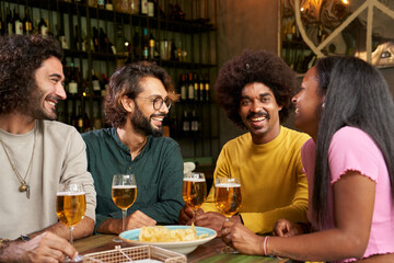 Group of young people drinking beer in a bar. This four multi-ethnic friends are smiling and having fun. Celebration of millennial colleagues enjoying together. Social gathering concept