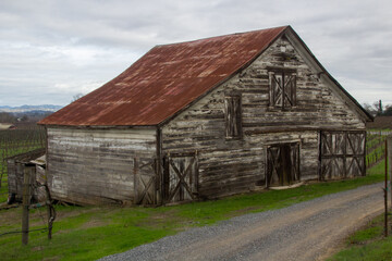 Fototapeta na wymiar An old weather worn wooden barn with a red metal roof stands beside a dirt road. Green grass is around the side of the barn. A cloudy sky is behind the barn