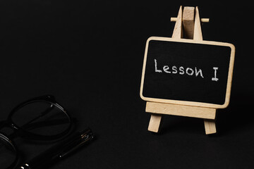 a small black writing board with "Lesson I" written on it, next to glasses and a pen on a black background (selective focus)