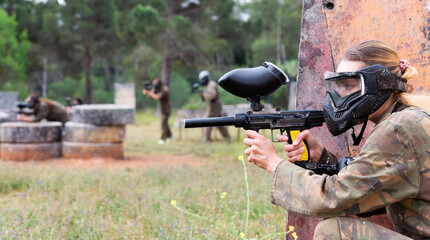Portrait of girl paintball player in mask who is aiming in opponents on paintball field