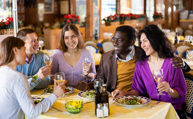 Multiracial group of happy adult people having fun in cozy restaurant during dinner, chatting and drinking wine..