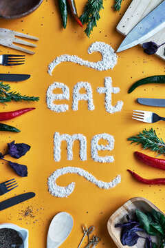 Top view of lettering Eat me made of potato starch in the middle of yellow table decorated with cutlery, herbs and pepper 