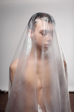 Conceptual shot of naked young man wrapped in plastic sheet standing and looking down sadly against white wall background 
