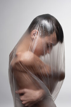 Side view of thin shirtless young man wrapped in polythene sheet embracing himself and looking down gloomily on white wall background 