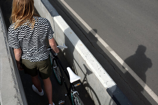 High angle rear view of man with long blond hair going along narrow sidewalk  with his bike while crossing the bridge 