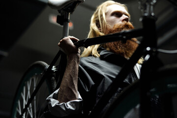 Low angle view of young long-haired man with full red beard carrying his bicycle with one hand while going through underground parking  