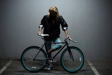 Young bearded man with long blond hair standing beside his retro bike with blue rims in underground garage and looking down    