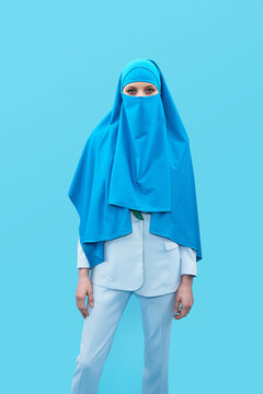 Portrait of unrecognizable woman in elegant suit and blue niqab standing against blue background and looking at camera  