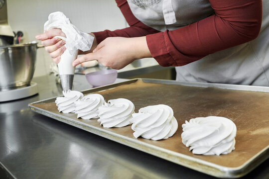 Unrecognizable female pastry chef using piping bag while making meringue nests in confectionery kitchen 