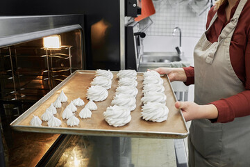 Unrecognizable female confectioner putting baking tray with meringue nests into the oven in restaurant kitchen 