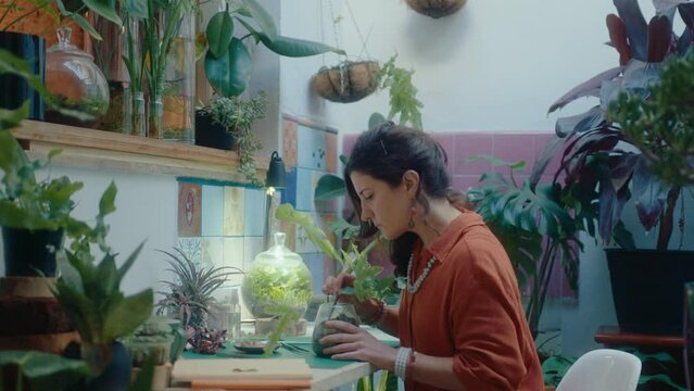 Female florist using tweezers to clean soil in flower pot and spraying plant leaves with water at the workplace in a houseplant shop. Medium shot, side view