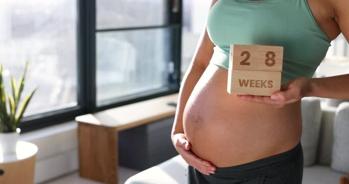 Pregnant woman showing 28 weeks pregnancy belly bump for maternity photoshoot holding wooden blocks sign.