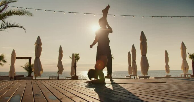 The guy helps his friend stand on his head on a special rug and supports his legs. Yoga classes and morning exercises together