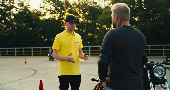 A male instructor in a yellow T-shirt and cap tells a biker with a beard about the rules of driving a motorcycle