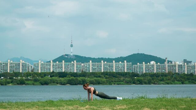 Sporty Woman Stretches Back and Train Core Doing Plank With Knee Tuck Exercises on Grassy Lawn in Han River Park on Summer Morning, N Seoul Tower on Namsan Mountain in Background