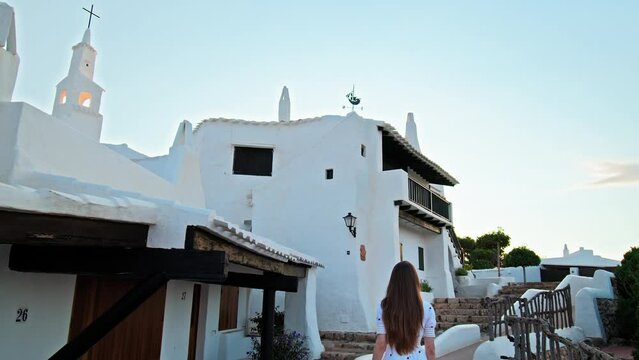 Beautiful young girl walking in Binibeca Coastal Village with whitewashed houses. A female enjoying relaxing tourism at Binibeca Vell a Calm Cove Resort in Menorca, Balearic Islands, Spain.