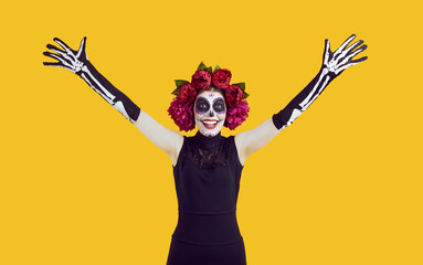 Positive woman - zombie with raised up hands isolated on yellow background. Santa muerte young lady with artistic halloween makeup on her face. Skeleton hands raised in greeting.