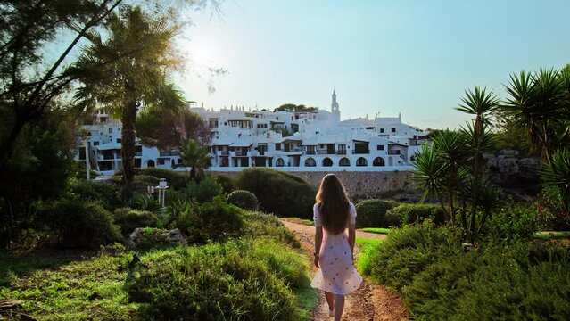 Beautiful girl walking amongst tropical nature towards Binibeca Vell in Menorca. A female tourist walks by green trees and nature on an early sunrise in a resort town in Balearic Islands, Spain.