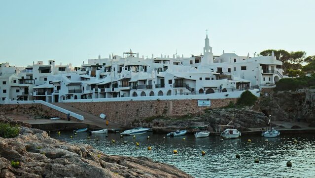 Boats by the seashore of Binibeca Vell with a town view in the background in 4K. Calming sea and early morning scenery in Menorca, Balearic Islands, Spain with whitewashed houses and rocky caves.