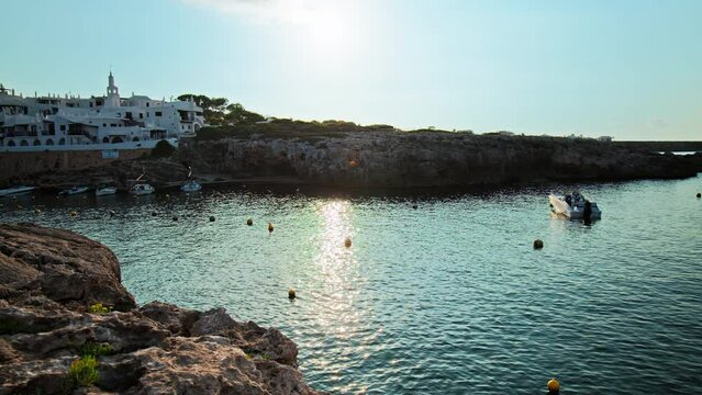 Boats by the seashore of Binibeca Vell with a town view in the background in 4K. Calming sea and early morning scenery in Menorca, Balearic Islands, Spain with whitewashed houses and rocky caves.