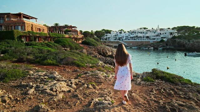 Beautiful girl in white dress long hair walking on a rocky seashore by the sea. Attractive female exploring beautiful nature early in the morning during sunrise in Binibeca Vell, Menorca, Spain