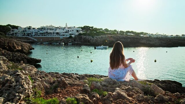 Girl in white dress long hair sitting on a rocky seashore with a beautiful view. Attractive female enjoying nature early in the morning during sunrise in Binibeca Vell, Menorca, Spain