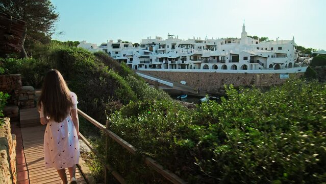 Beautiful sunrise over a seashore in Menorca with a wooden sidewalk and nature. Woman walking on a footpath pavement made of wood by the sea in Binibeca Vell, Balearic Islands, Spain.