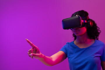 Biracial woman using vr headset and pointing on neon pink to purple background