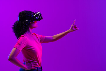 Biracial woman using vr headset and pointing on neon pink to purple background
