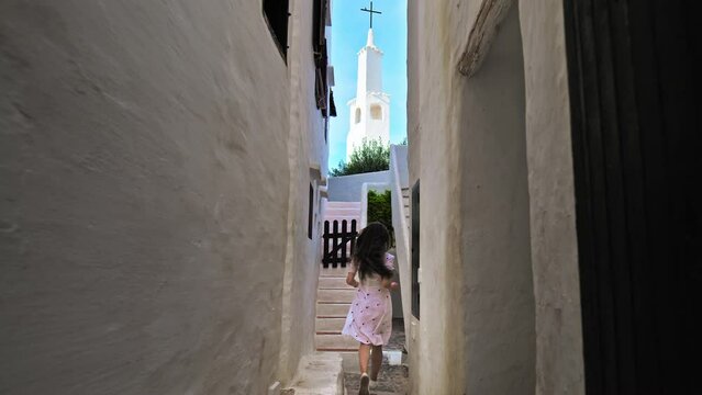 Beautiful young girl in a white dress running in slow motion in narrow streets. A woman with long hair running by whitewashed houses in Binibeca Vell, Menorca, Balearic islands, Spain.