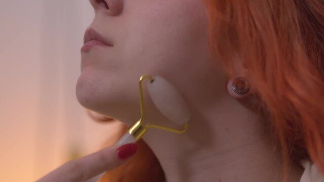 Fixed shot of a girl using a jade roller on her face with closed eyes. She had red hair. Left face profile framing