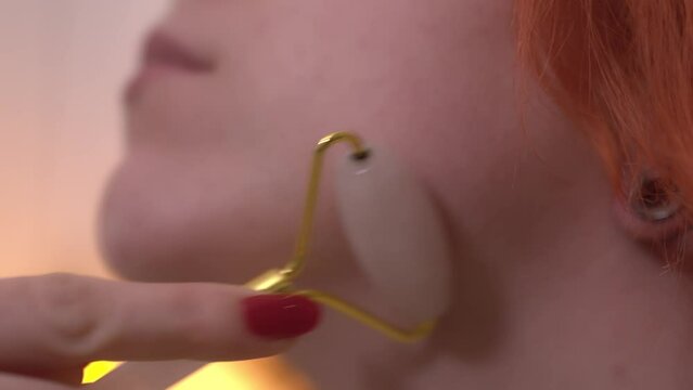 Cose up shot of a girl using a jade roller on her face. She had red hair. Left face profile framing. The jade roller tool is in a golden color